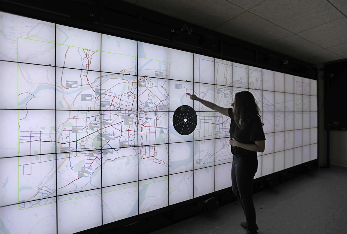 Grazia, seen in profile, pointing at one spot on a wall-sized display made of dozens of screens, showing a city's network on a map.