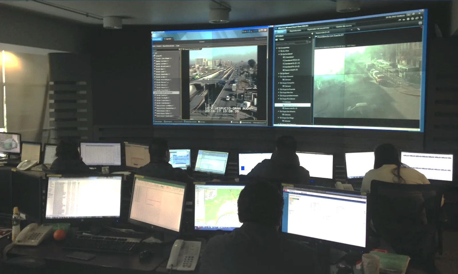 Operators, seen from behind, at desks with many monitors, under a video wall showing traffic cameras.